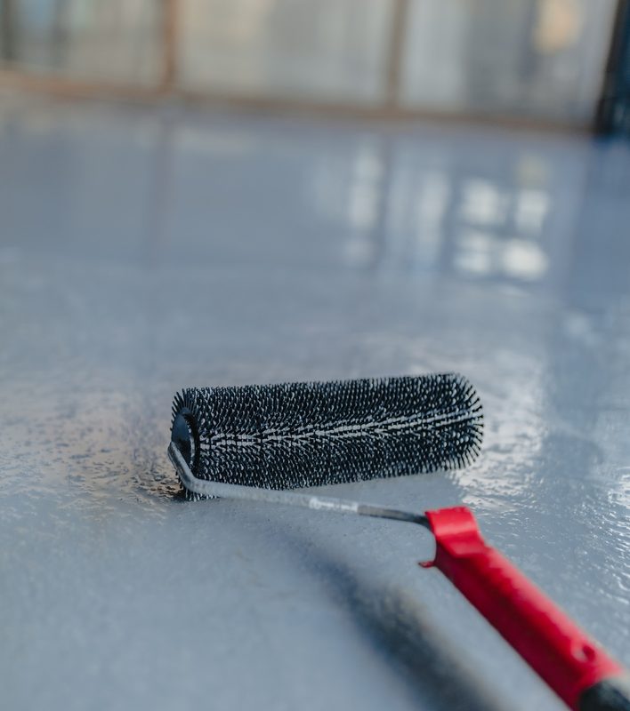 Squeegee pulling a light-colored floor coating across gray concrete in a sunlit room